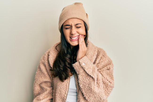 Young hispanic woman wearing wool sweater and winter hat touching mouth with hand with painful expression