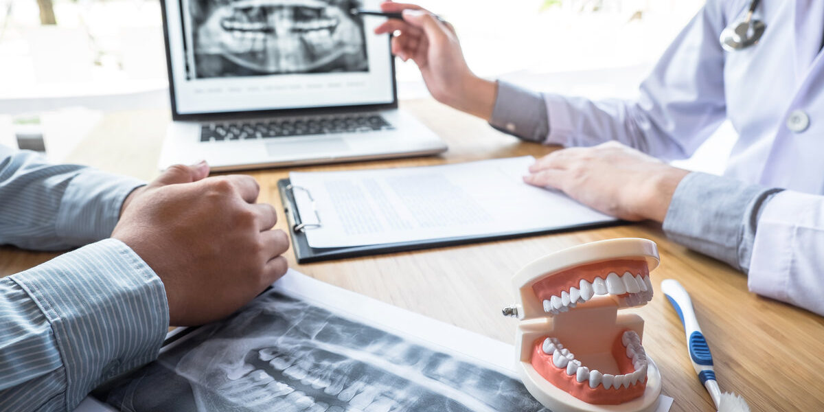 Professional Dentist showing jaw and teeth the x-ray photograph and discussing during explaining the consultation treatment issues with patient and writing history list on report.