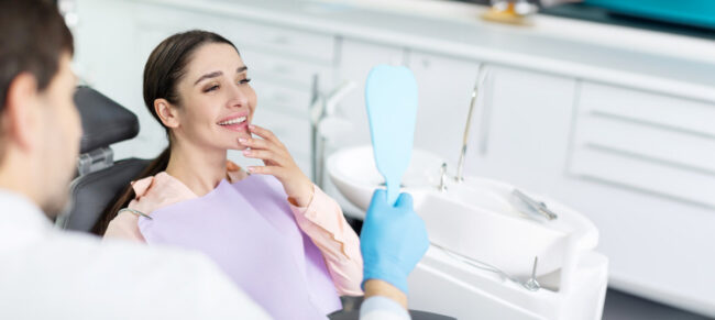 Smiling woman looking in mirror after dental procedure in clinic, enjoying result of teeth whitening result Smiling woman looking in mirror after dental procedure in clinic, enjoying result of teeth whitening result
