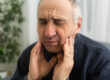Old man with toothache. Elderly senior man has toothache. Unhappy man face in tooth pain sitting on sofa at home, feel sick unwell. Sad aged man hand holding his chin. Adult suffering toothache