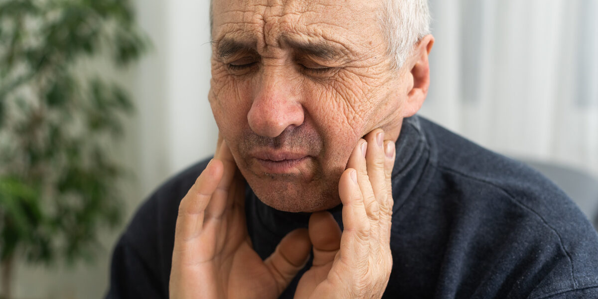 Old man with toothache. Elderly senior man has toothache. Unhappy man face in tooth pain sitting on sofa at home, feel sick unwell. Sad aged man hand holding his chin. Adult suffering toothache