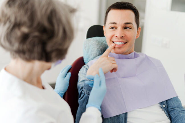 Male patient sitting in dental chair pointing at teeth, talking with professional dentist doctor Young attractive male patient sitting in dental chair pointing at teeth, having pain, talking with professional dentist doctor in modern dental clinic. Concept of dental care, health care